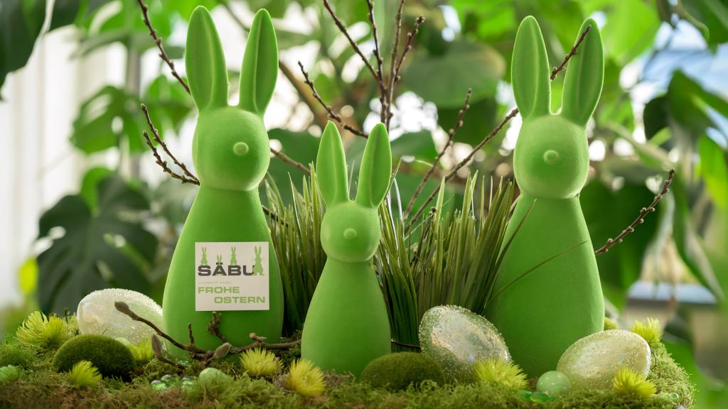 Drei grüne Osterhasen stehen inmitten grünen blättern, Gras und davor liegen Ostereier