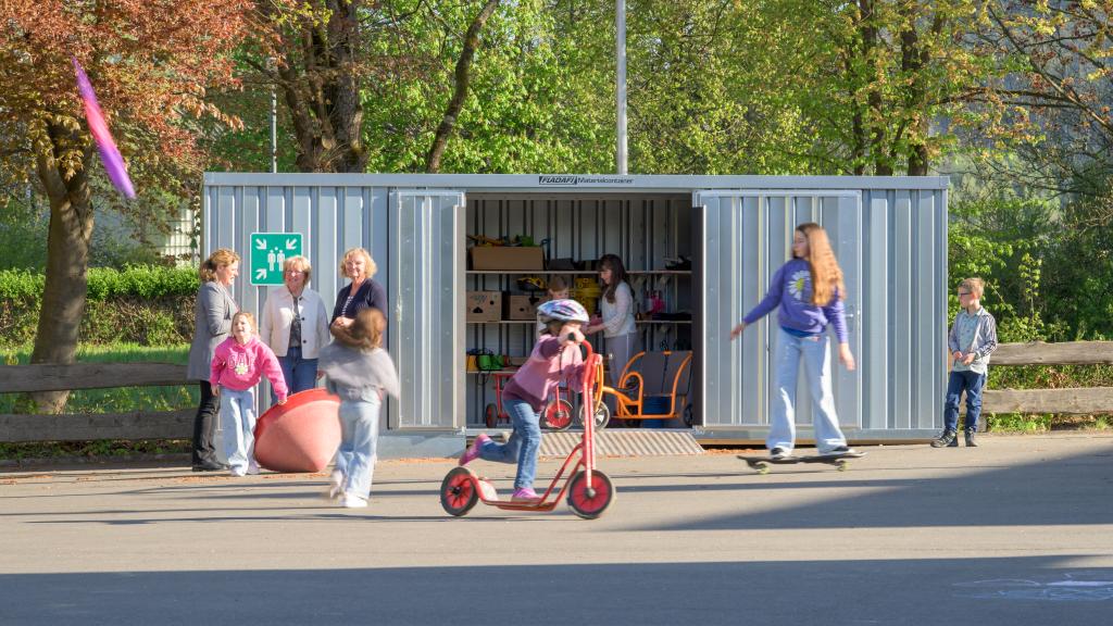 Außenansicht des Materialcontainers der Grundschule Dattenfeld mit spielenden Kindern auf dem Schulhof im Vordergrund.
