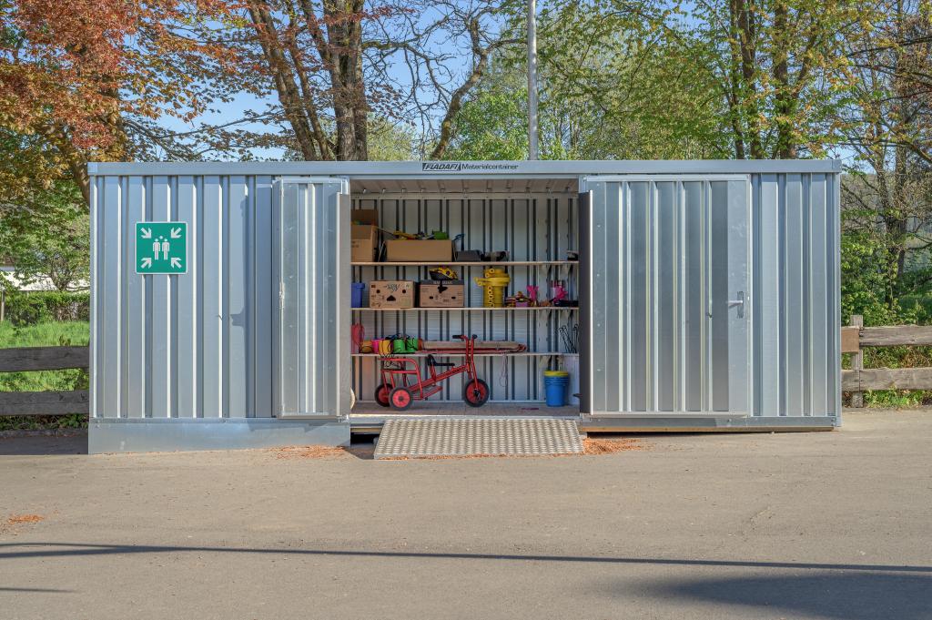Außenansicht unseres Materialcontainers mit Doppelflügeltüren. Im Inneren ist die funktionale Zubehör gut sichtbar darunter verschiedenes Spielzeug für den Einsatz auf dem Schulhof.
